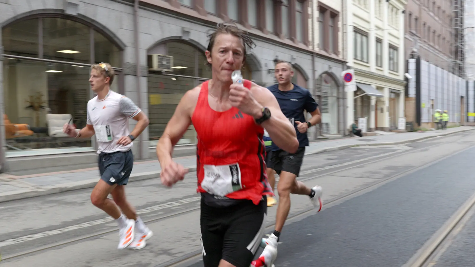 Matt Smith løper i Oslo Maraton 2026 med en gel i hånda, i rød singlet i en gate i sentrum